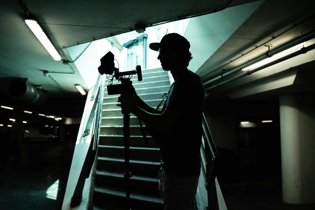 pexels-photo-6728042-6728042 Silhouette of a filmmaker holding a camera in an underground parking area with stairs.